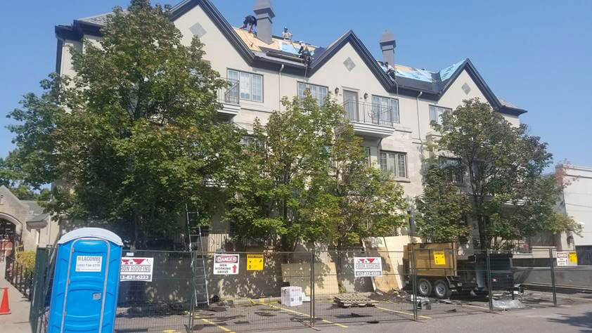 A large apartment building undergoing roof repairs, with workers on the roof, warning signs and fences below, and a blue portable toilet in front—showcasing expert roofing for businesses in Ottawa. Trees partially obscure the building on a clear, sunny day.