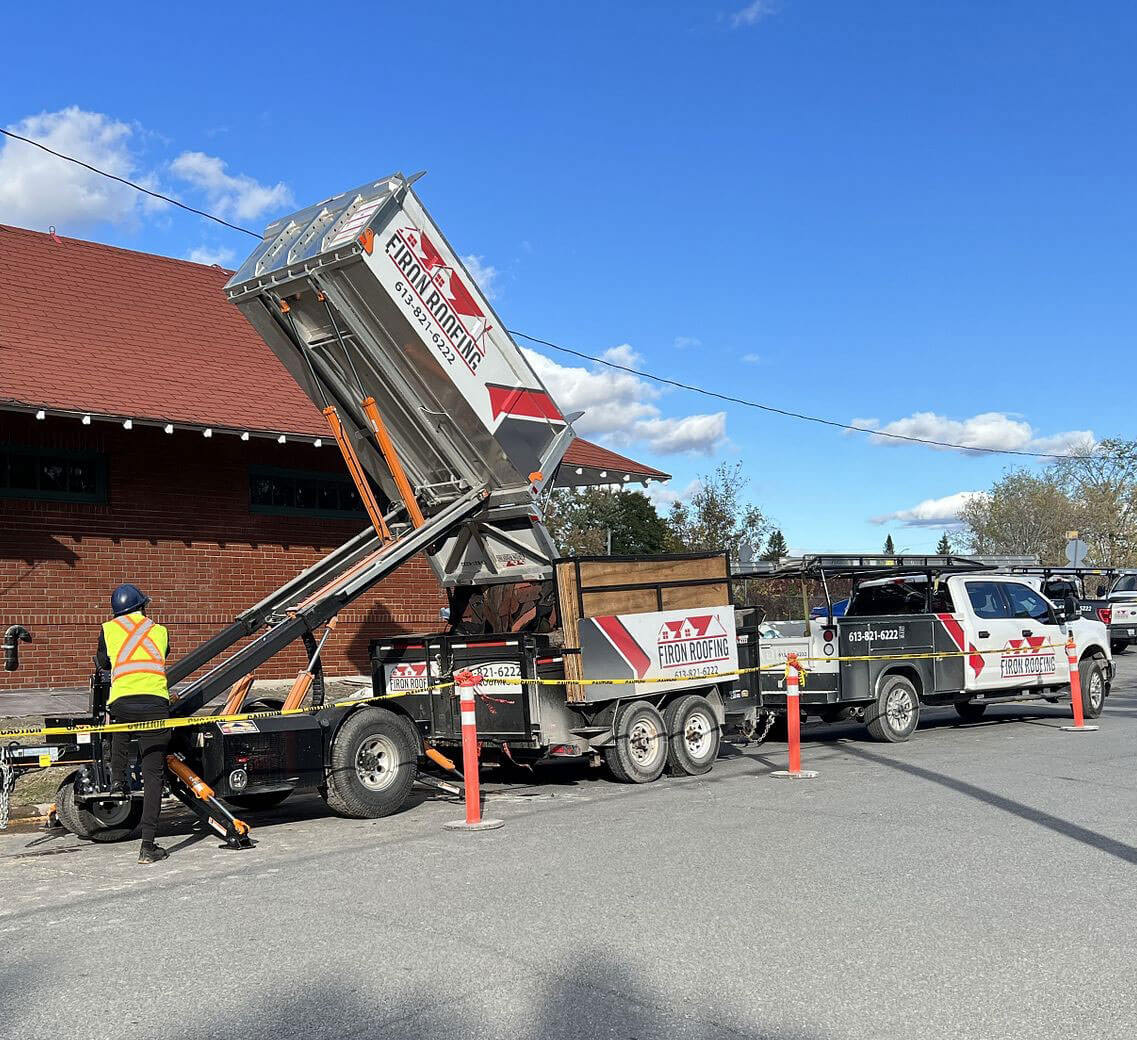 A worker in a safety vest and helmet stands by a dump trailer labeled Fincham Paving, unloading materials next to a company truck on a sunny street beside a brick building. Orange cones mark the work area—Firon Roofing for Ottawa businesses visible nearby.