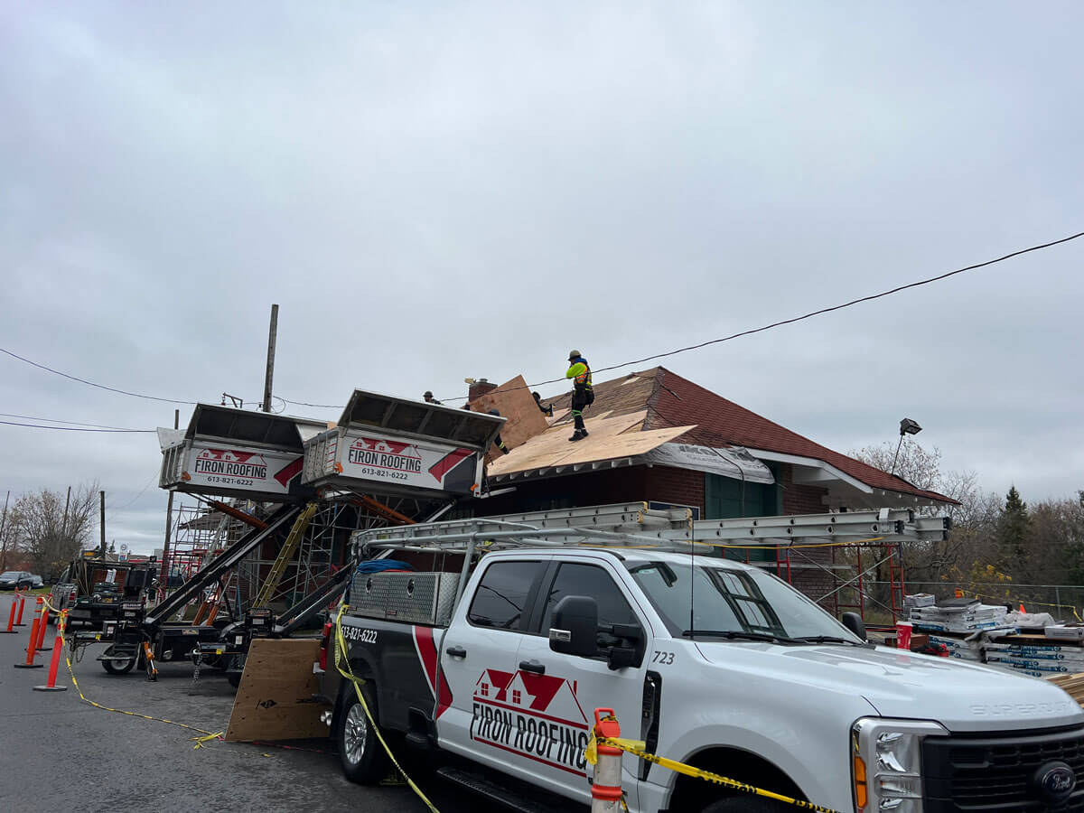 A roofer in safety gear works on the roof of a building under construction, with two Ferron Roofing trucks and construction equipment on site under a cloudy sky. Yellow caution tape surrounds this commercial roofing Ottawa project.