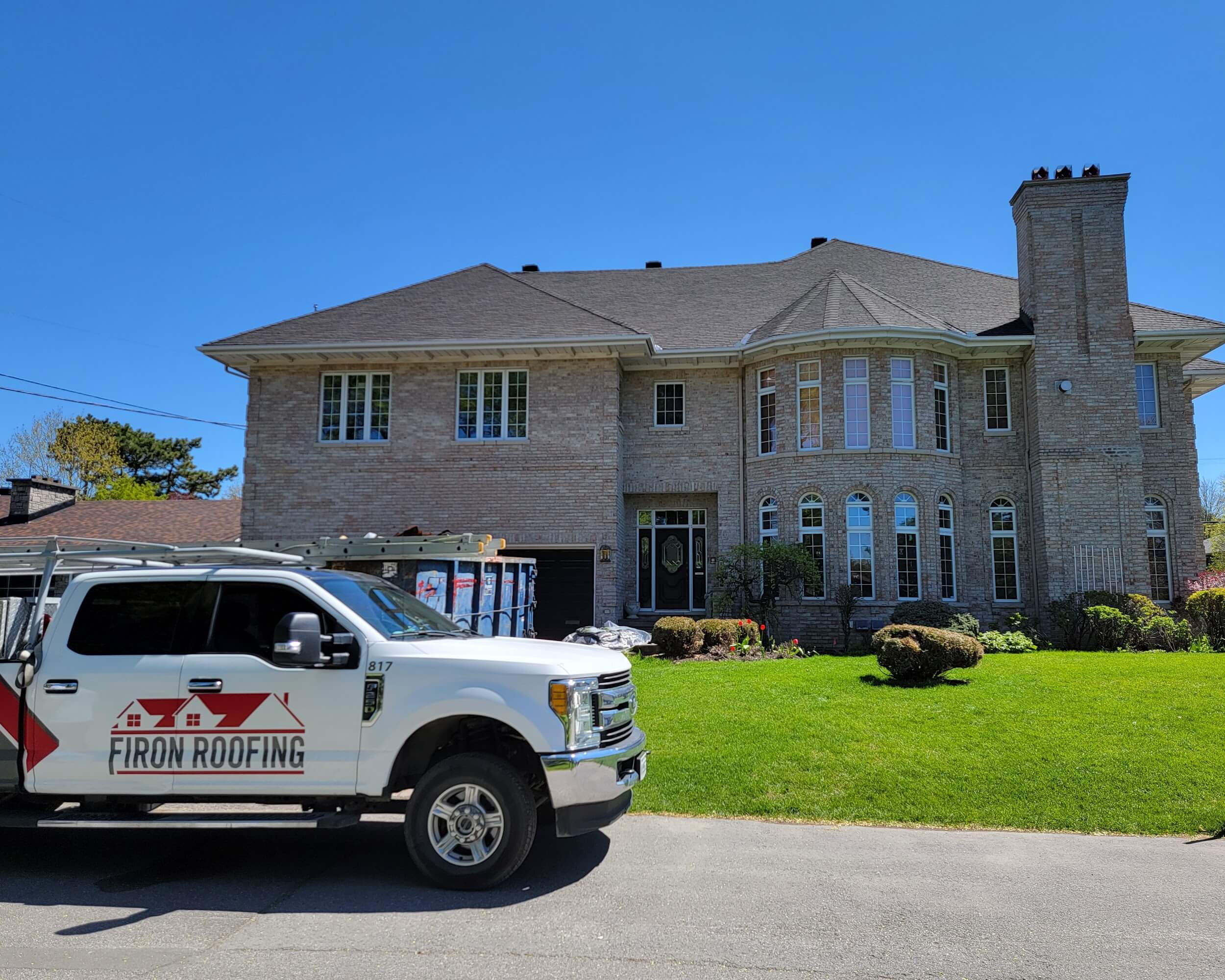 A white Firon Roofing truck, serving Ottawa / Gatineau, is parked in front of a large, two-story brick house with a dark roof and tall windows on a sunny day. A dumpster and some equipment are visible in the driveway.