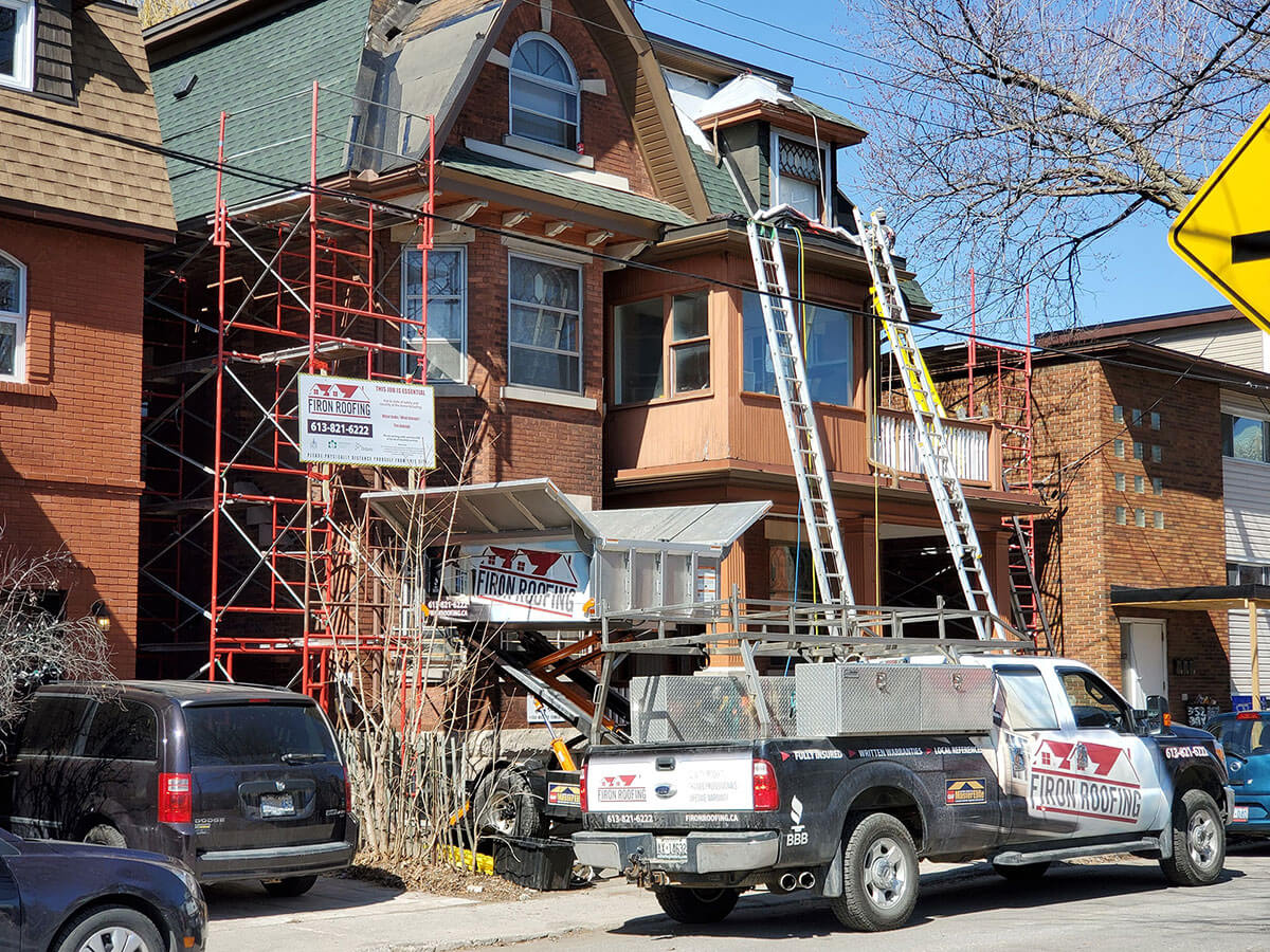 A house undergoing roofing work with scaffolding and ladders set up. A pickup truck with Adam Roofing signage is parked in front, along with roofing materials and other vehicles parked along the street.