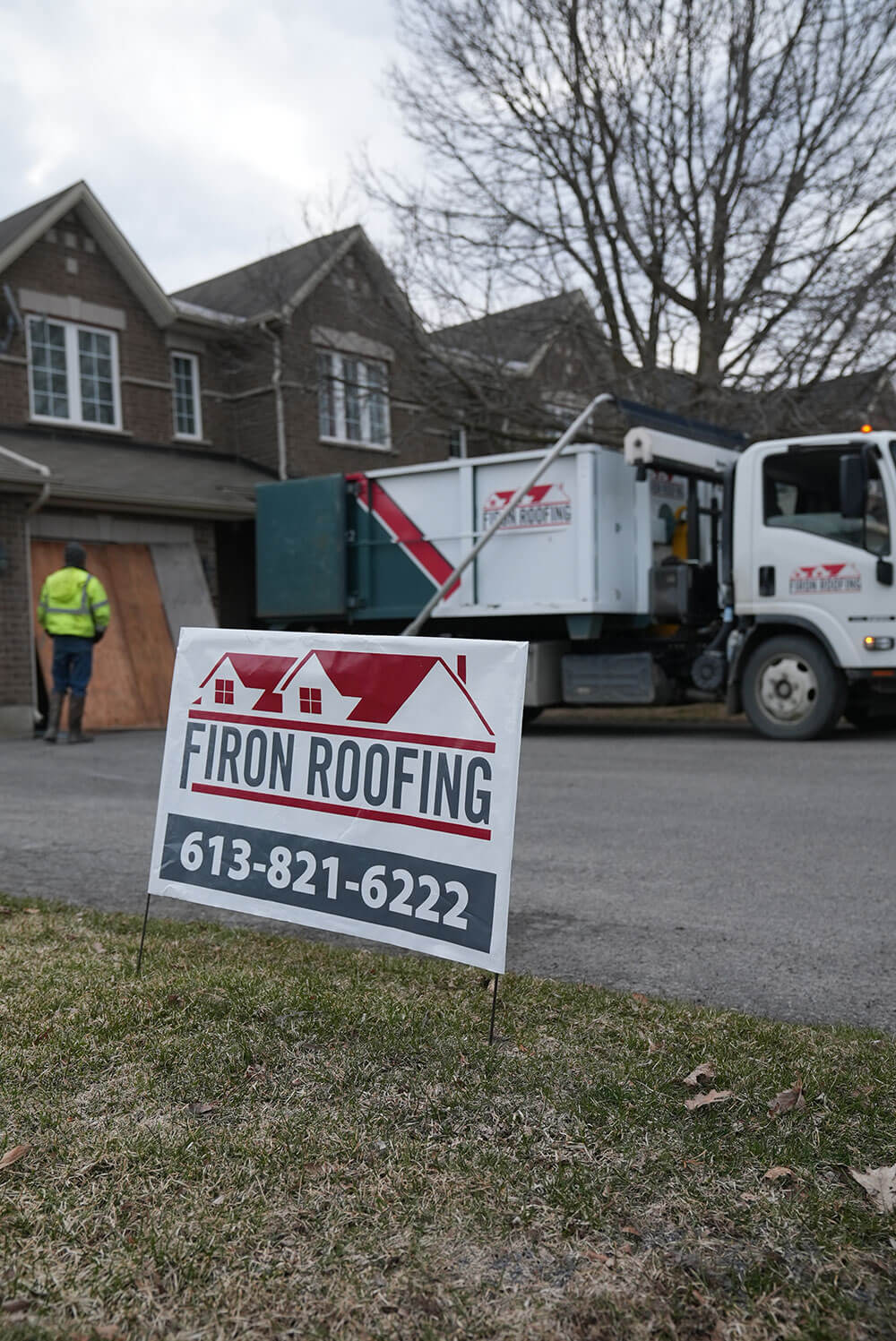 A lawn sign for Firon Roofing with a phone number is in the foreground. In the background, a worker stands near a house and a Firon Roofing truck is parked by the curb. Trees without leaves are visible.