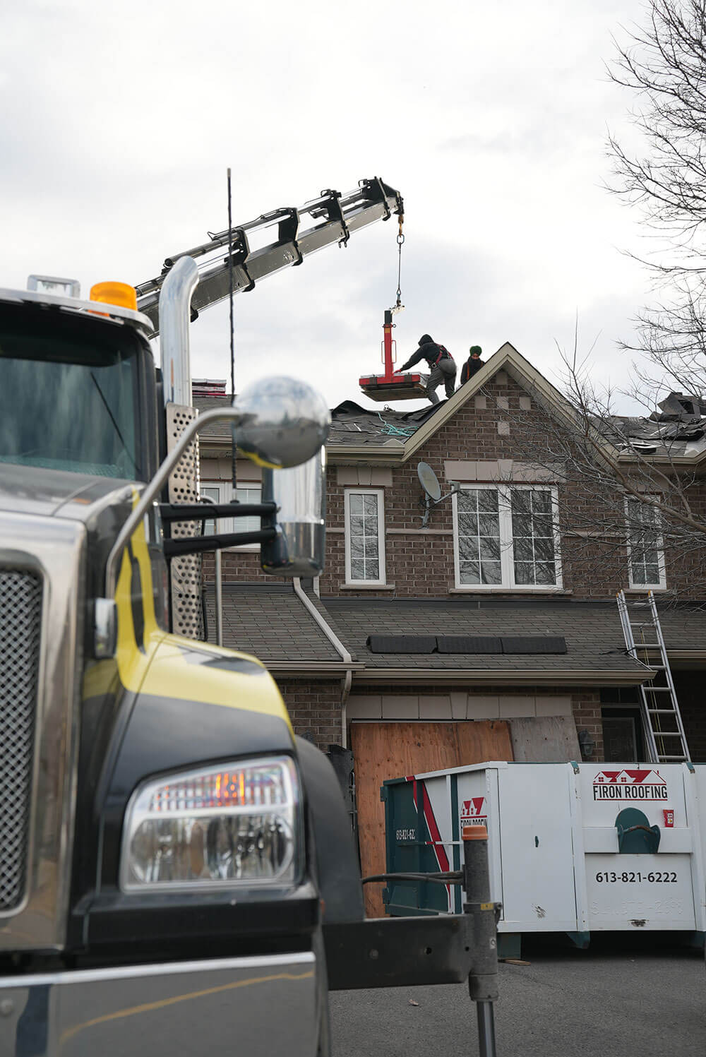 A crane truck lifts roofing materials to workers on the roof of a two-story house in Ottawa. A ladder leans against the house, and a trailer with a roofing company logo sits in the driveway, highlighting expert work on roofs in Ottawa.