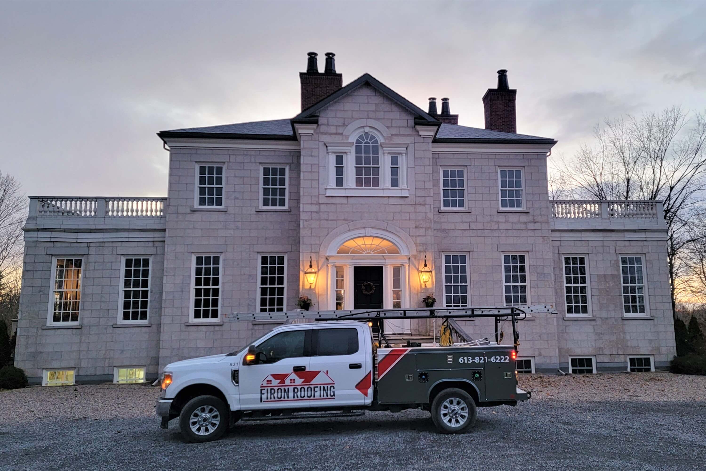 A white truck with Firon Roofing branding is parked in front of a large, symmetrical stone house with tall windows and chimneys, showcasing expert roofing Ottawa services as exterior lights glow at dusk.