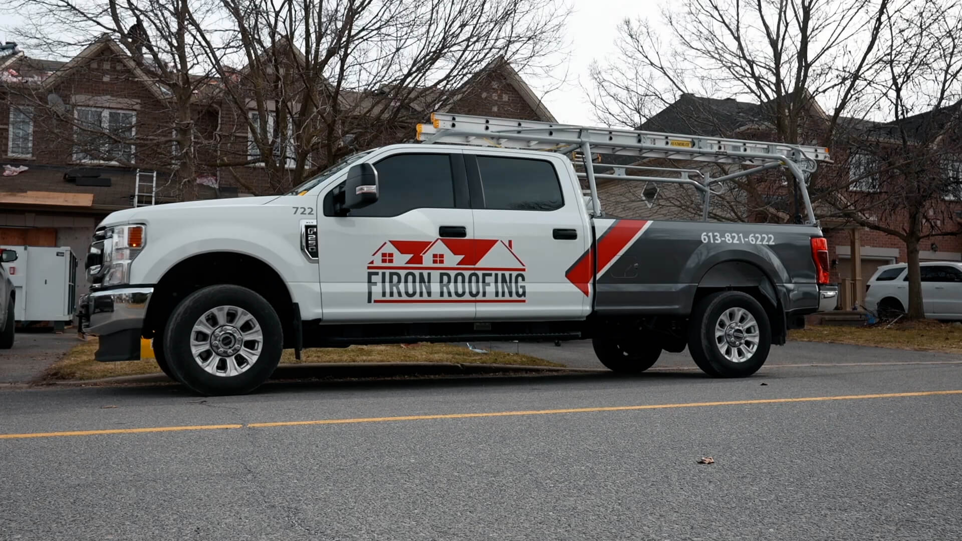 A white pickup truck with Firon Roofing and a red roof logo is parked on a suburban street, ready for roofing in Nepean. A ladder rests on the truck’s rack, with houses and bare trees visible in the background.