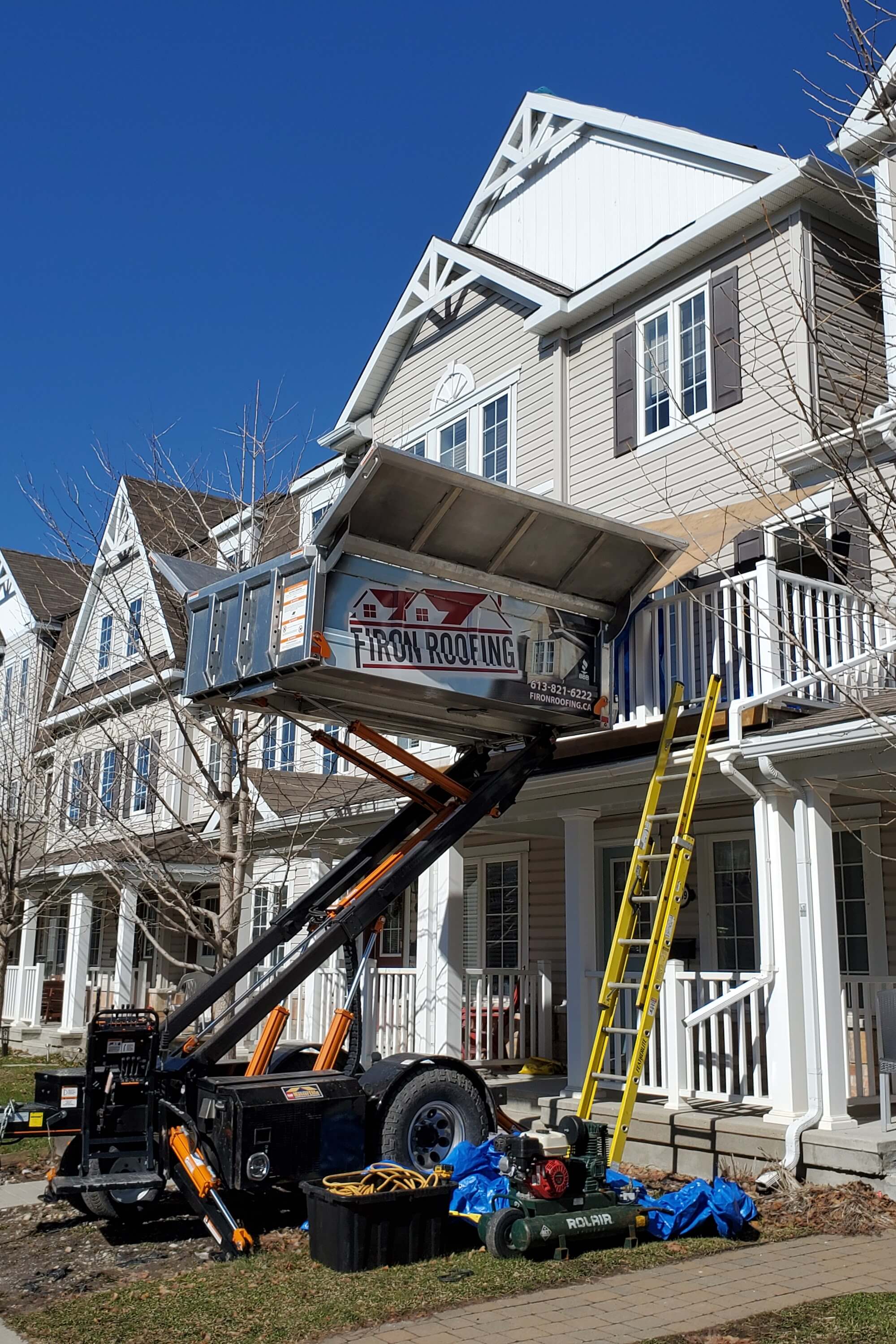 A roofing debris container is lifted by a hydraulic trailer to a second-story balcony during a roof replacement. A yellow ladder leans against the townhouse, with tools and tarps spread across the lawn below.