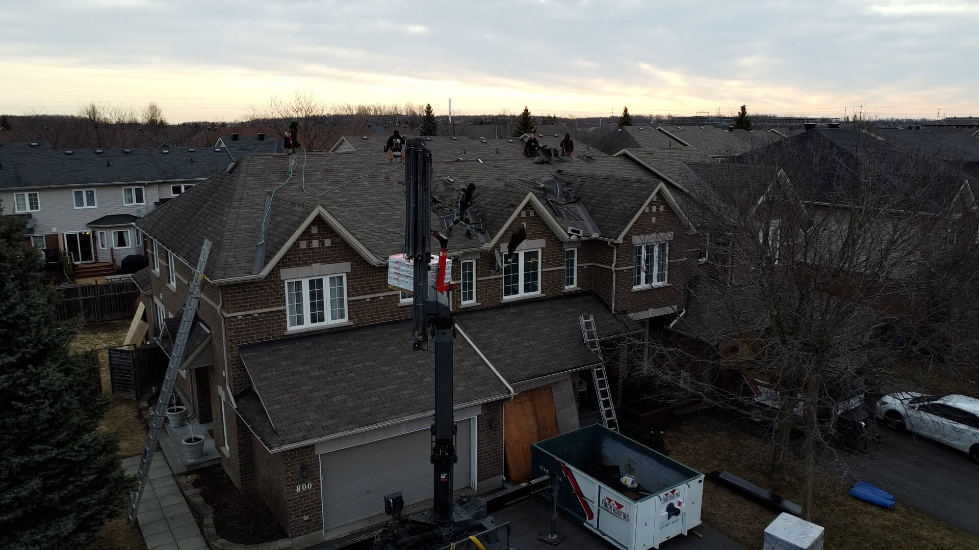 A team of workers performs a roof replacement, repairing or installing shingles on a two-story suburban house using ladders and a crane, with equipment and materials on the ground below. The sky is overcast at sunset.