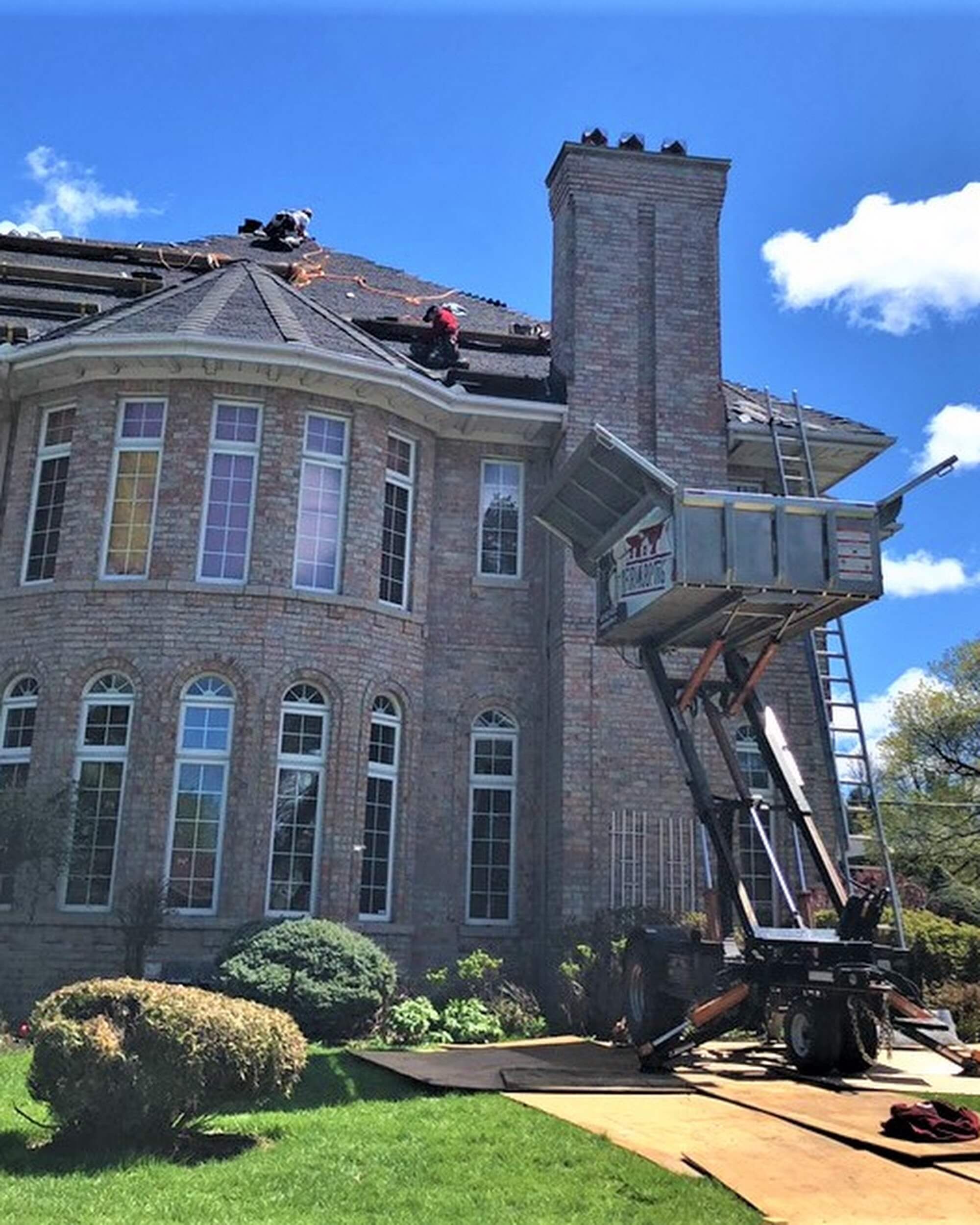 Workers are providing roofing services on a large brick house, using a lift to transport materials. The house features tall windows and a manicured lawn under a partly cloudy sky.