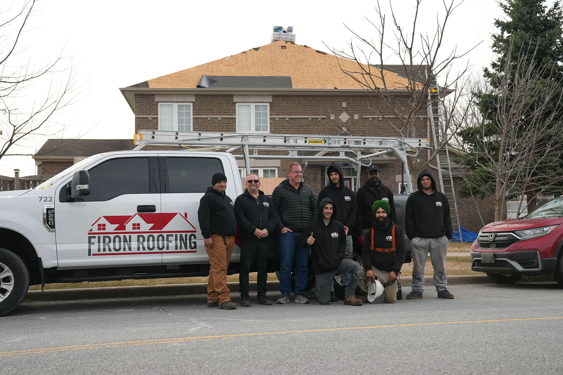 A group of eight people in work clothes, part of the Firon Roofing team, stand in front of a Firon Roofing truck with a house under construction and a ladder in the background. Some are smiling and posing for the photo.