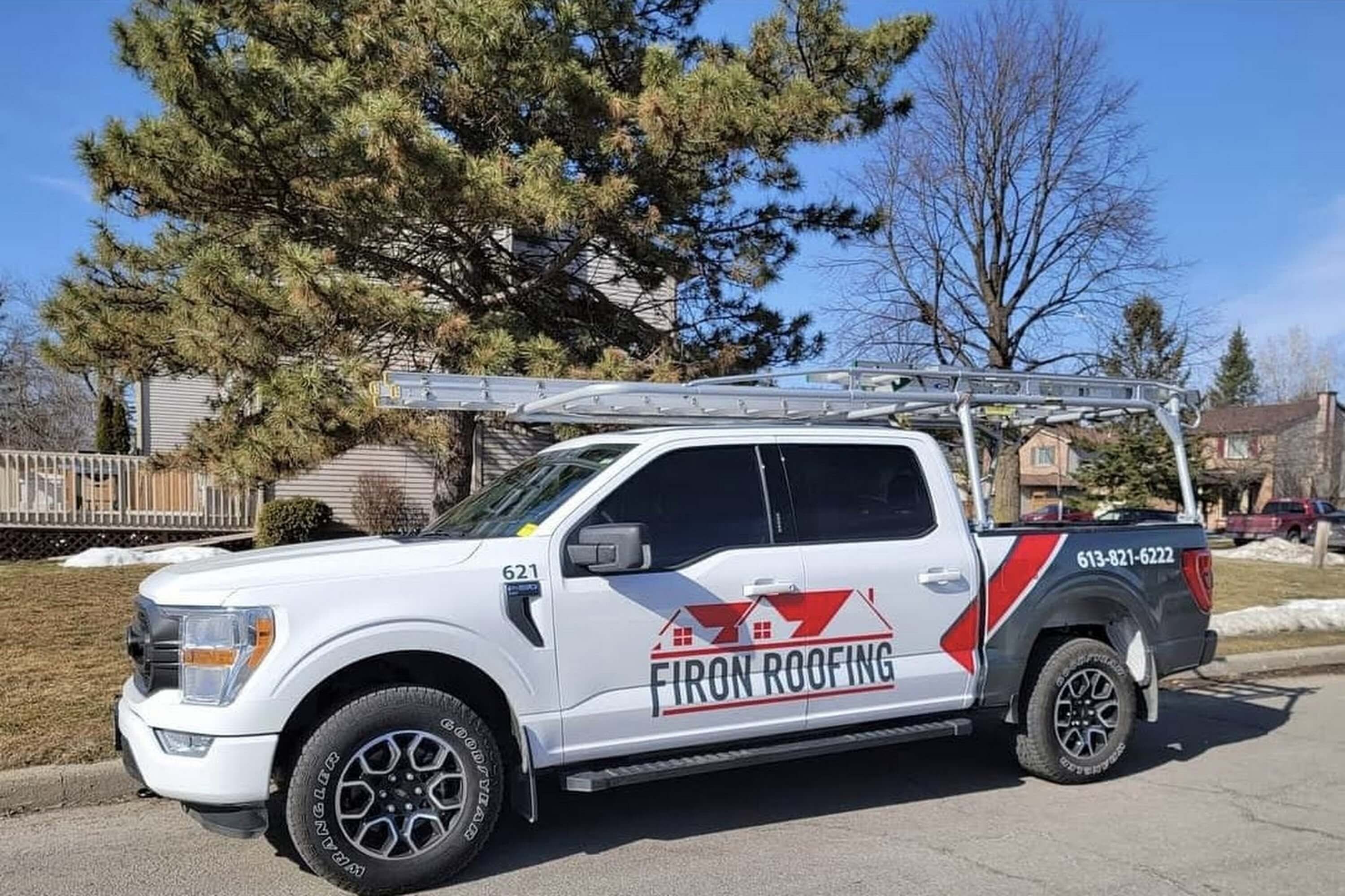 A white pickup truck with a ladder rack and ladder on top is parked on a residential street. The truck displays Firon Roofing, an Ottawa roofing company, with a phone number and images of red roofs on the side.
