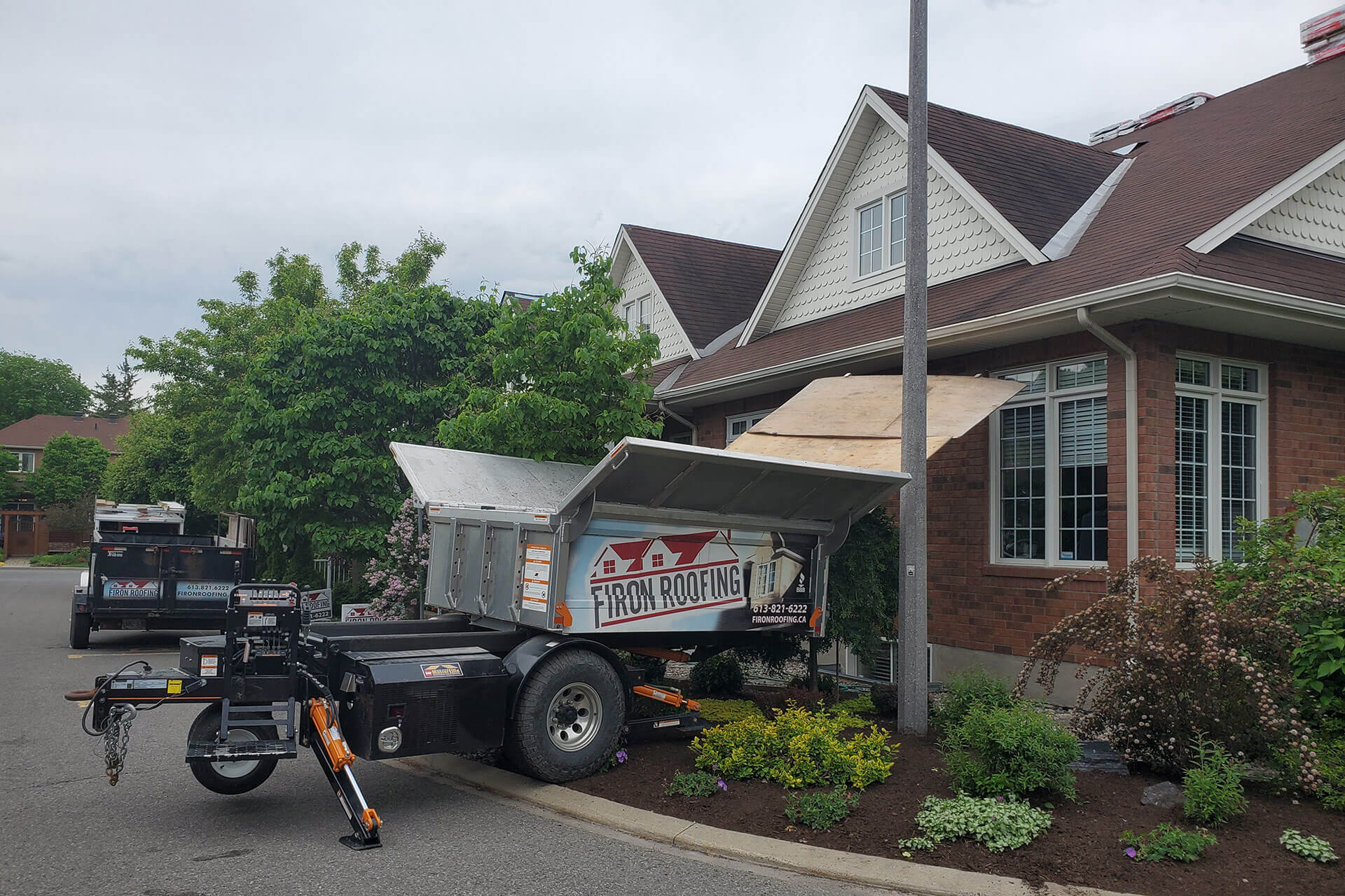 A trailer labeled From Roofing is parked beside a brick house. A wooden chute extends from the roof to the trailer, likely for roofing shingles cleanup. Trees, bushes, and other landscaping surround the house.