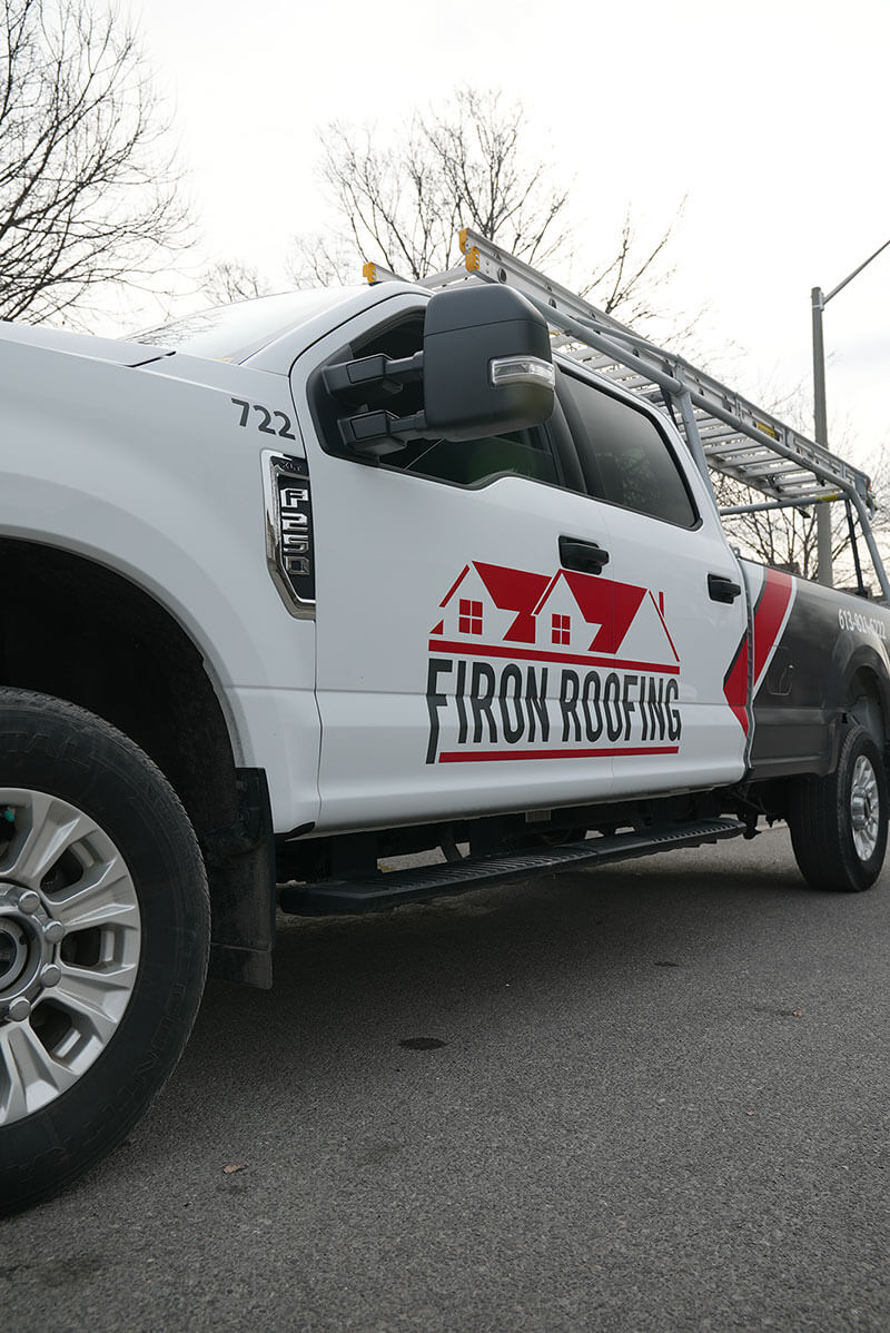 White pickup truck with a ladder on top parked on a street. The truck has Firon Roofing, a roofing company serving the Ottawa and Gatineau region, written on the door with a red house roof logo. Trees and a cloudy sky are visible in the background.