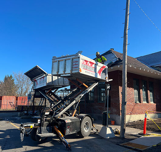 A pickup truck labeled “From Roofing” is parked in front of a brick building. Workers use a cherry picker to access the sloped roof, where roofing for business Ottawa repairs are underway. Leafless trees surround the scene.