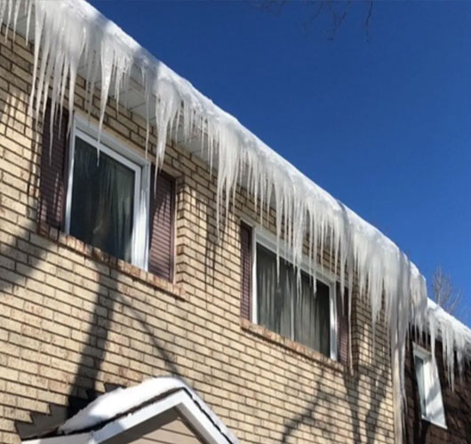 A two-story brick house with long, sharp icicles hanging from the roof and closed blinds on the windows under a clear blue sky. Snow covers the lower roof, highlighting the need for timely roof snow and ice removal.