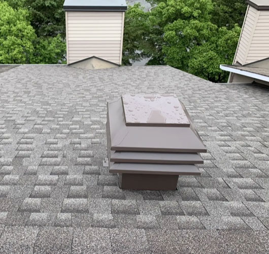 A brown roof vent, part of recent roof ventilation upgrades, sits on a gray shingled roof dotted with water droplets. In the background, trees and neighboring buildings with beige siding and similar vents are visible.