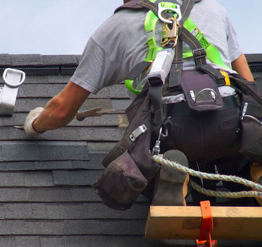 A worker wearing safety gear and a harness installs asphalt shingles during roof repairs in Gatineau, using a tool to secure them. A safety rope is attached for fall protection.