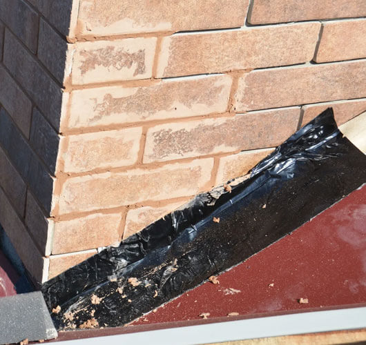 Damaged roof flashing next to a brick chimney, with black tape used as a temporary roof flashing repair where the roof and chimney meet. The brickwork and roofing material are clearly visible.