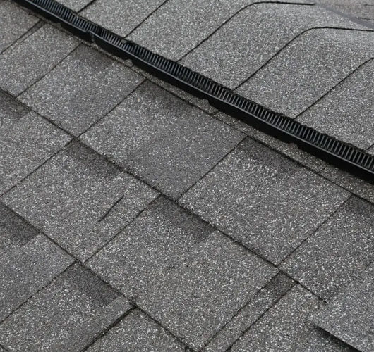 Close-up view of gray asphalt roof shingles with a black ridge vent running along the peak, highlighting the texture and overlapping pattern while illustrating effective roof ventilation.