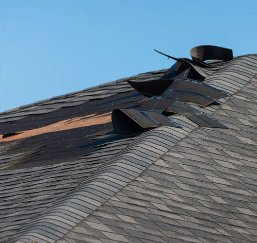 Damaged roof with several asphalt shingles peeled and lifted, exposing the wooden structure underneath, against a clear blue sky. The roof wind damage is evident as the shingles appear to be lifted and in need of repair.
