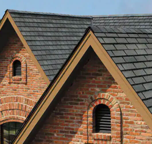 Two gabled rooftops with gray designer shingles and red brick exterior walls, each featuring an arched louvered vent. The architectural style suggests a classic or traditional home design.