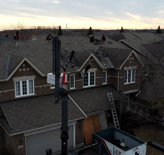 A group of workers performs a roof replacement on a two-story brick house, using a crane to lift materials. Ladders and a dumpster are visible below in the suburban neighborhood at dawn or dusk.