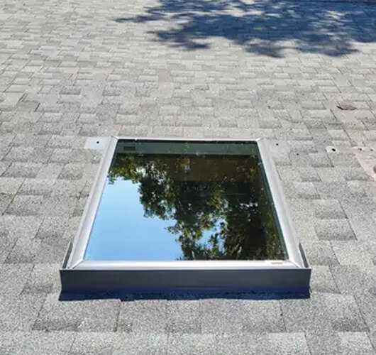 A rectangular skylight set into a gray shingled roof reflects the blue sky and tree branches above, showcasing the natural beauty and quality of skylights Ottawa homeowners appreciate.