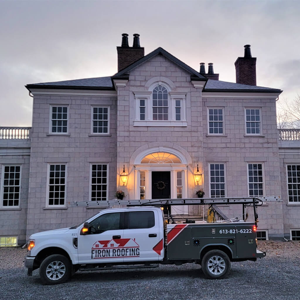 A Firon Roofing truck, showcasing roofing Ottawa branding and a ladder, is parked in front of a large, elegant stone house at dusk. Warm lanterns glow by the entrance, highlighting expert care and attention to detail.