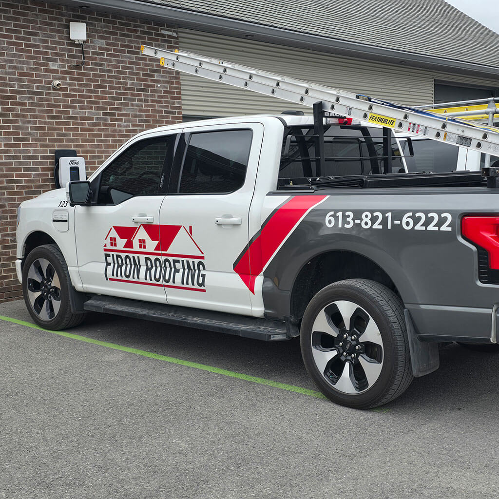 A gray and white pickup truck with Firon Roofing and a phone number printed on the side, parked outside a brick building with ladders in the bed, ready to handle any roof repair Ottawa / Gatineau clients may need.