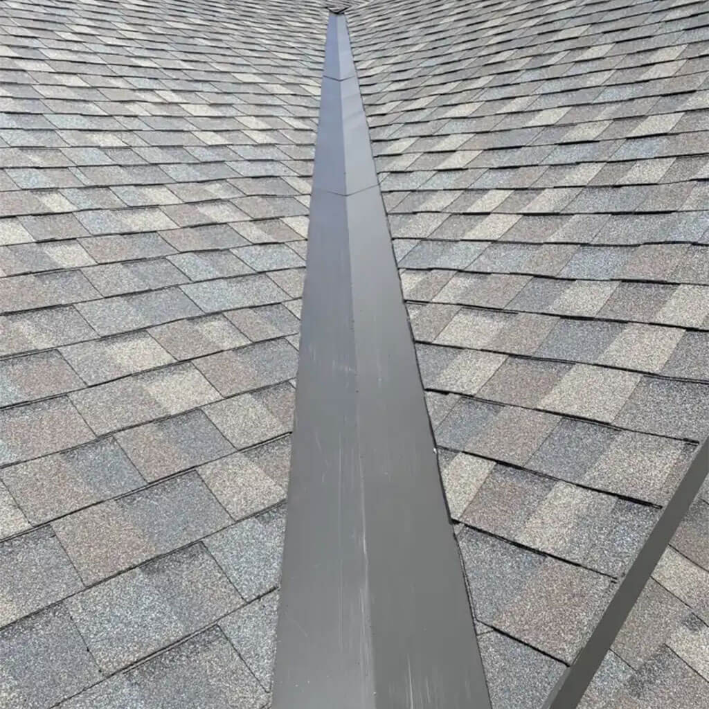 A close-up view of a shingled rooftop, showing a metal valley running down the center where two roof slopes meet. The shingles are gray with slight color variations.