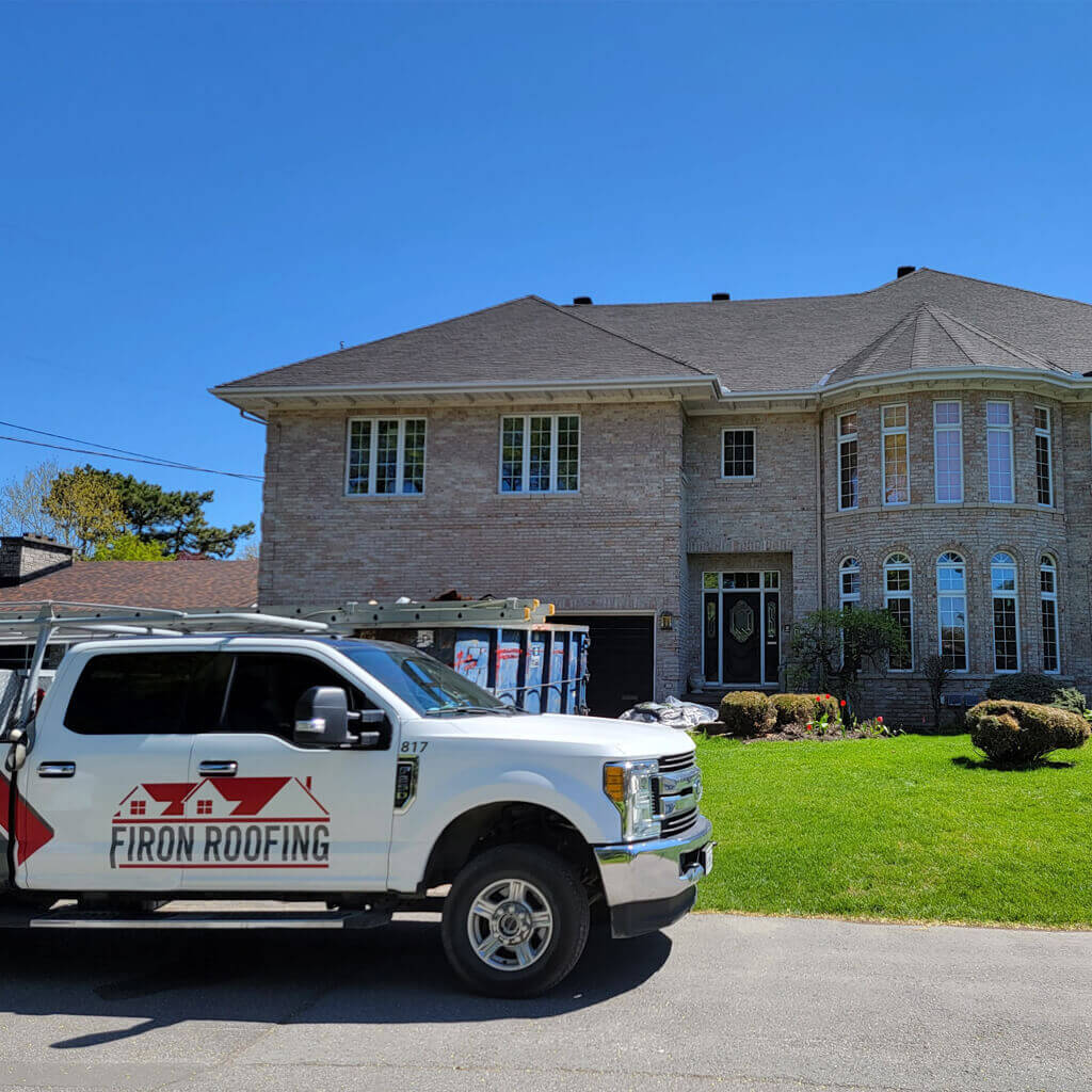 A white Firon Roofing truck, serving Ottawa / Gatineau, is parked in front of a large, two-story brick house with a dark roof and tall windows on a sunny day. A dumpster and some equipment are visible in the driveway.