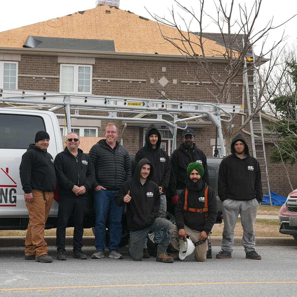 A group of eight people, including skilled roofing installers Ottawa, poses in front of a Firon Roofing truck on a suburban street, with a house under construction behind them. Some wear work clothes and hoodies; the atmosphere is casual and friendly.